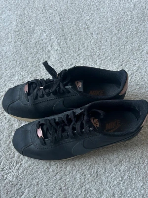 Svarta Nike sneakers med rosa detaljer - Snygga svarta Nike sneakers med diskreta rosa detaljer på tungan och hälen. Skorna har klassisk låg profil, svart snörning och sula i gummi. Perfekta för dig som gillar stilrena och sportiga skor med en twist. Säljer åt min mamma, passar ca 40-41❤️