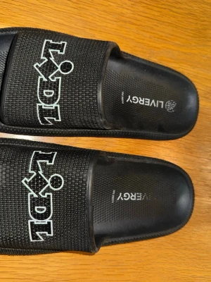 Svarta Lidl badtofflor från Livergy - Svarta sandaler från Livergy med stort vitt och blått Lidl-tryck på ovandelen. Tillverkade i syntetmaterial med räfflad sula och öppen tå. Perfekta för chill dagar eller stranden.