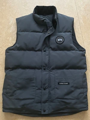 Svart dunväst från Canada Goose - Snygg svart dunväst från Canada Goose med hög krage och två stora framfickor. Västen har ett klassiskt Canada Goose-märke på bröstet och stängs med tryckknappar. Perfekt för lager-på-lager och håller dig varm under kyliga dagar.