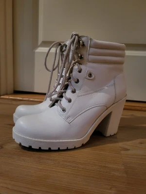 Vita chunky stövletter med snörning - Snygga vita stövletter med chunky klack och grov sula. Skorna har snörning framtill, vadderad kant och metallöljetter. Tillverkade i ett läderliknande material som ger en cool och modern look. Perfekta för dig som vill sticka ut.