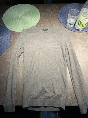 Grå stickad tröja från New Look - Säljer en stilren grå stickad tröja från New Look Men. Tröjan har rund halsringning, långa ärmar och ribbade muddar vid ärmslut och nederkant. Perfekt basplagg för lager på lager och enkel att matcha med olika outfits.