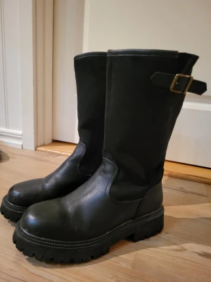 Svarta chunky stövlar med spänne - Säljer ett par svarta höga stövlar med grov sula och dekorativt spänne upptill. Stövlarna har en robust design och är tillverkade i ett slätt, syntetiskt material. Perfekta för en edgy och trendig look.
