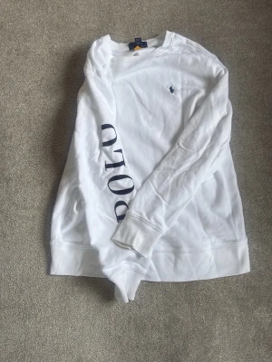 Vit sweatshirt från Polo Ralph Lauren - Vit sweatshirt med blå Polo-tryck längs ena sidan och klassiska Polo Ralph Lauren-loggan broderad på bröstet. 