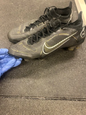 Nike Mercurial svart/guld fotbollsskor - Snygga Nike Mercurial fotbollsskor i svart med guldiga detaljer och vit swoosh på sidan. Skorna har snörning, låg profil och dobbar för spel på gräs. Tillverkade i syntetmaterial för lätt vikt och bra bollkontroll. Perfekta för dig som vill sticka ut på planen.