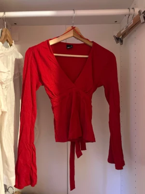Röd omlottopp med knyt - Trendig röd omlottopp med långa ärmar och knyt i midjan. Från Gina tricot, storlek xs. Djup v-ringning framtill och mjukt, lätt material som ger en snygg passform. Perfekt att styla med jeans eller kjol för en cool look. Denna topp finns även i 4 andra färger. Skriv om ni är intresserade 