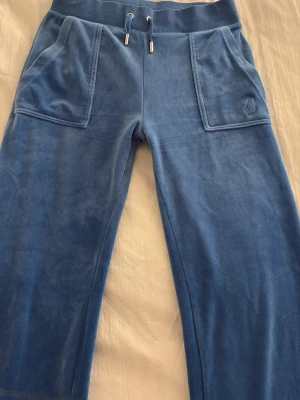 Blå velour mjukisbyxor Juicy Couture - Blå mjukisbyxor i mjuk velour från Juicy Couture. Byxorna har bred resår i midjan med snörning, stora fickor fram och bak med knappdetaljer samt broderad logga på ena fickan. Perfekta för en chill och avslappnad stil.
