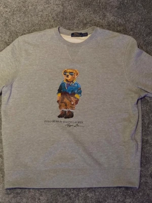 Grå sweatshirt Polo Bear Ralph Lauren - Tjena! Säljer nu min gråa sweatshirt från Ralph Lauren med ikoniskt Polo Bear-tryck på bröstet, använd endast ett fåtal gånger. Tröjan har rund halsringning, långa ärmar och ribbade muddar. Priset är inte ristat i sten och kan diskuteras vid en krånglar affär! Hör av dig vid några funderingar.