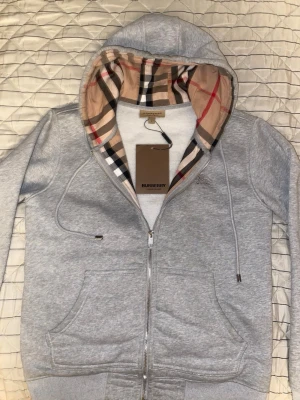 Grå Burberry zip - Snygg grå zip hoodie från Burberry med den klassiska rutiga mönstrade huvan i beige, svart, rött och vitt. Hoodien har dragsko i huvan, två fickor fram och ribbade muddar. Tillverkad i mjukt bomullsmaterial och har en exklusiv känsla. Perfekt för en stilren och avslappnad look.