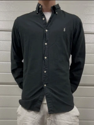 Svart skjorta från Ralph Lauren - Klassisk svart skjorta från Ralph Lauren med button-down krage och broderad logga på bröstet. Skjortan har långa ärmar, vita knappar och är tillverkad i mjuk bomull. Perfekt till jeans eller chinos för en clean look.