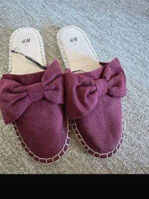 Vinröda tofflor med rosett från H&M - Säljer ett par vinröda tofflor från H&M med stora dekorativa rosetter framtill. Tofflorna har öppen häl, mjuk textil på ovansidan och kontrastsömmar runt sulan. Perfekta för att chilla hemma i stil.