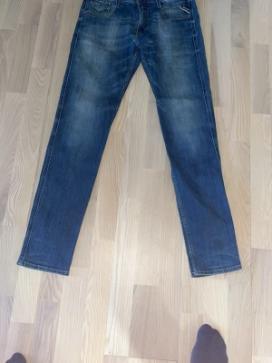 Blå raka jeans från Replay - Säljer ett par klassiska blå jeans från Replay med rak passform och snygga slitningar. Jeansen har fem fickor, gylf med knapp och dragkedja samt detaljerade sömmar. Perfekta för en avslappnad och trendig stil.