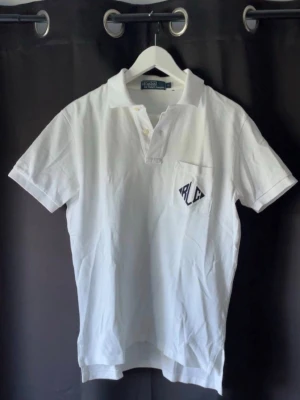 Vit pikétröja från Ralph Lauren - Skick 10/10, Klassisk vit pikétröja från Ralph Lauren med korta ärmar och krage. Tröjan har knappslå framtill och en liten bröstficka med RL-logga i mörkblått. Tillverkad i mjuk bomull som känns skön mot huden. Perfekt till jeans eller shorts.