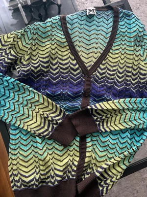 M Missoni zigzag-mönstrad kofta - Färgstark kofta från M Missoni med klassiskt zigzag-mönster i turkos, limegrönt, blått och svart. Koftan har bruna ribbade muddar, v-ringning och knäppning framtill. Materialet är tunt och glittrigt, perfekt för att lyfta vilken outfit som helst.
