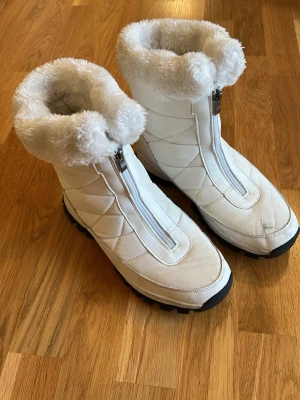 Vita vinterstövlar med päls - Säljer ett par vita vinterstövlar med fluffig fuskpäls runt skaftet och dragkedja framtill. Stövlarna har grov sula för bra grepp och quiltad design på sidorna. Perfekta för kalla dagar när du vill hålla dig varm och snygg. Vet ej märket