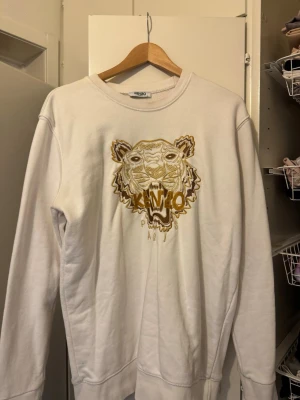 Kenzo sweatshirt storlek L - Säljer en vit sweatshirt från Kenzo med det ikoniska tigerhuvudet broderat i guld och brunt på bröstet. Tröjan har rund halsringning och ribbade muddar vid ärmslut och nederkant. Perfekt statement-plagg för dig som gillar streetwear och snygga detaljer.