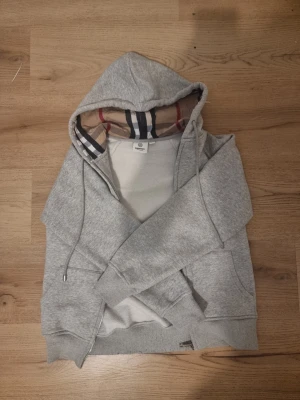  Burberry - Säljer en grå zip hoodie från Burberry med klassiskt rutigt foder i huvan och dragsko. Hoodien har fickor framtill och ribbade muddar vid ärmar och nederkant. Perfekt för dig som gillar stilrena och exklusiva plagg.