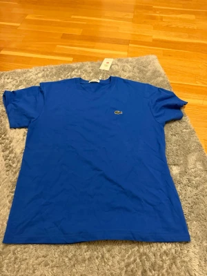 Blå t-shirt från Lacoste - Klassisk blå t-shirt från Lacoste med den ikoniska krokodilloggan broderad på bröstet. T-shirten har rund halsringning och korta ärmar. Tillverkad i mjuk bomull som känns skön mot huden. Perfekt för en avslappnad och stilren look.