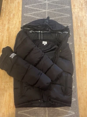 Svart pufferjacka från burberry - Snygg svart pufferjacka från Björn Borg med huva och dragkedja framtill. Jackan har två fickor med dragkedja och en logga på ärmen. Perfekt för kalla dagar med sin vadderade design och sportiga look.