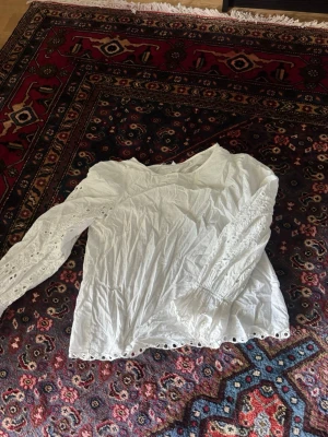 Vit blus med broderade detaljer - Söt vit blus med långa ärmar och broderade spetsdetaljer längs ärmar och nederkant. Blusen har rund halsringning och är tillverkad i ett lätt bomullstyg. Perfekt för en romantisk och bohemisk stil. Super fin 