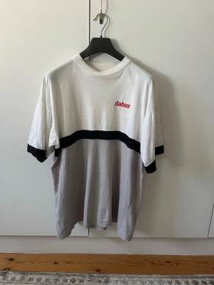 Vit och grå bomull t-shirt från Dabus strl XL - Säljer en t-shirt från Dabus i vitt och ljusgrått med svart rand över bröstet och ärmsluten. T-shirten har rund halsringning. Tröjan har lite gul färgning vid halsen (se sista bilden för mer förtydligande). Storlek XL (syns ej på bilden men står XL på lappen). Material 100% bomull.