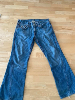 Blå bootcut jeans från True Religion - Säljer ett par klassiska blå bootcut jeans från True Religion med ikoniska sömmar och fickor med lock bak. Jeansen har låg midja, fem fickor och är gjorda i slitstark denim. Perfekta för dig som gillar en avslappnad men ändå trendig look.