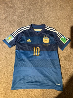 Argentina matchtröja Adidas blå - Säljer en blå Argentina matchtröja från Adidas med nummer 10 i guld, officiella VM 2014-detaljer och AFA-emblem på bröstet. Tröjan har vita ränder på axlarna, korta ärmar och är tillverkad i lätt funktionsmaterial. Perfekt för fotbollsfans!