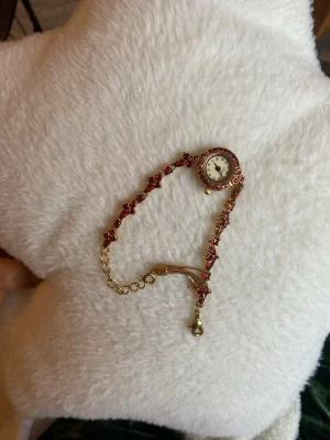 Guldig guldpläterad  armbandsklocka med zirikon stenar - Unik armbandsklocka i guldton med röda vinröda stenar. Klockan har ett runt ur med dekorativa detaljer runt boetten och ett smyckat armband med blommönster. Perfekt för dig som vill ha något extra till din stil.