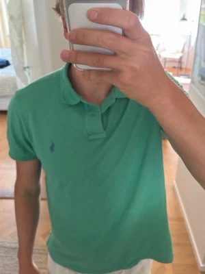 Grön pikétröja från Ralph Lauren - Snygg grön pikétröja från Ralph Lauren med klassisk krage och korta ärmar. Tröjan har två knappar vid halsen och den ikoniska broderade loggan i blått på bröstet. Perfekt för en avslappnad och stilren look. Storlek L men passar M och L. Modellen på bilden är 190 cm lång.