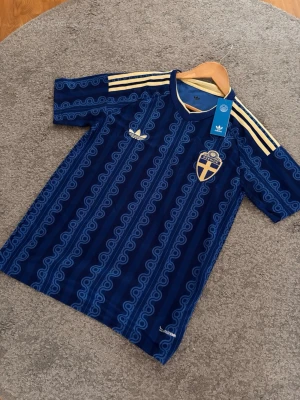 Blå Sverige t-shirt från Adidas - Blå t-shirt från Adidas med Sveriges landslagsemblem på bröstet och gula detaljer. Tröjan har korta ärmar, mönstrad design och klassiska Adidas-ränder på axlarna. Perfekt för dig som vill visa ditt stöd för Sverige. Skriv gärna för fler bilder eller info!🙌🏼🇸🇪.                             OBS: den är cirka 2 cm kortare än vanlig S då den är uppsyrd.