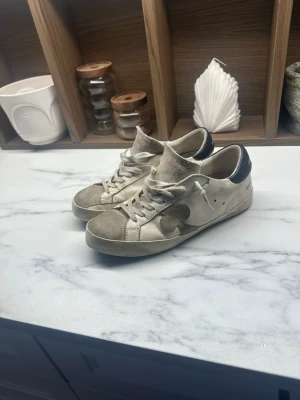Beige sneakers från The Gallerist - Snygga beige/vitta  sneakers från The Gallerist med svart hälparti och råa kanter på plösen. Skorna har en unik detalj med en mörk fläck på sidan och text på hälen. Tillverkade i mocka och läder med vita snören och låg profil. Färgerna är klassiska och modellen är snygg och populär, pris kan diskuteras 