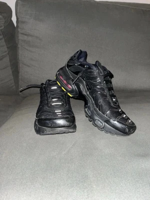 Nike Air Max Plus svarta sneakers - Säljer ett par Nike Air Max Plus sneakers i helsvart med mesh och syntetdetaljer. Skorna har synlig Air Max-dämpning i sulan, gula och rosa detaljer samt reflekterande inslag på ovandelen. Klassisk TN-logga på plösen och robust yttersula.