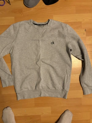 Grå sweatshirt från Calvin Klein - Snygg grå sweatshirt från Calvin Klein med klassisk rund halsringning och svart 'ck'-brodyr på bröstet. Tröjan har ribbade muddar vid ärmslut och nederkant, perfekt för en clean och stilren look.
