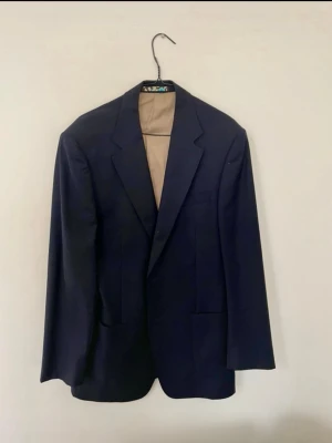 Loro Piana (56, L-XL) - Stilren mörkblå kavaj ifrån Loro Piana sydd i Italien i super 120s ull. Den har två knappar framtill och klassiska slag. Kavajen har raka fickor och ljus innerfoder. Passar perfekt till en dressad look och är enkel att matcha med skjorta eller t-shirt. 