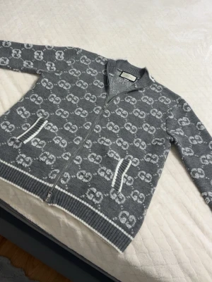 Grå ziptröja med GG-mönster från Gucci - Snygg grå ziptröja från Gucci med vitt GG-monogram över hela plagget. Tröjan har ribbade muddar, två fickor framtill med vita detaljer och dragkedja hela vägen. Perfekt för dig som gillar exklusiv streetwear och vill sticka ut.