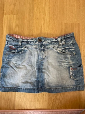 Blå jeanskjol med fickor - Säljer en blå jeanskjol med slitningar och flera fickor fram och bak. Kjolen har bälteshällor, knappstängning och en liten ficka med knappdetaljer på sidan. Perfekt för en avslappnad och cool stil.