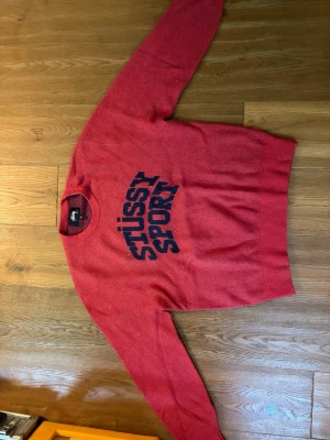 Röd Stüssy Sport tröja - Röd tröja från Stüssy Sport med blå logotyp på bröstet. Klassisk rund halsringning och ribbade muddar vid ärmslut och nederkant. Perfekt för dig som gillar streetwear och vill ha en snygg och bekväm tröja.
