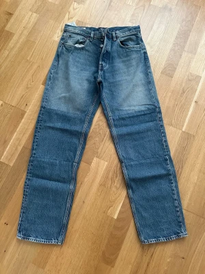 Blå raka jeans med läderpatch - Säljer ett par klassiska blå jeans med rak passform och fem fickor. Jeansen har en snygg brun läderpatch på bakfickan och är gjorda i slitstark denim. Perfekta för dig som gillar en tidlös och avslappnad look.