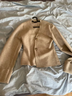 Beige cardigan från Gina tricot med knappar - Stilren beige cardigan från Gina tricot med två stora knappar framtill och v-ringad hals. Koftan har vida ärmar med dekorativ knappdetalj vid ärmslut och är gjord i ett mjukt, stickat material. Perfekt för lager på lager och enkel att matcha. Säljer pga att den inte kommer till användning använt 1 gång.