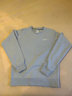 Ljusblå stickad tröja från Nike - Ljusblå stickad tröja från Nike med klassisk rund halsringning och lång ärm. Tröjan har en broderad vit Nike-logga på bröstet och ribbade muddar vid ärmslut och nederkant. Perfekt för en avslappnad och sportig stil.