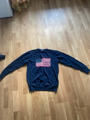 Mörkblå stickad Polo Club tröja med USA-flagga - Snygg mörkblå stickad tröja från Polo Club med en stor amerikansk flagga broderad på bröstet. Tröjan har rund halsringning och långa ärmar. Perfekt för dig som gillar klassisk preppy stil och vill sticka ut lite extra.