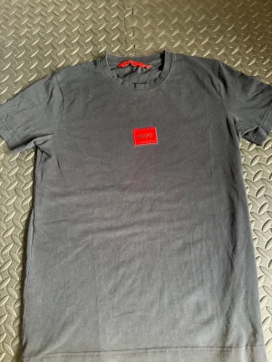 Svart t-shirt från Hugo med röd logga - Svart t-shirt från Hugo med klassisk passform och rund hals. På bröstet sitter en röd patch med HUGO-broderi som ger en clean och modern vibe. Tillverkad i mjuk bomull som känns skön mot huden. Perfekt till jeans eller shorts.