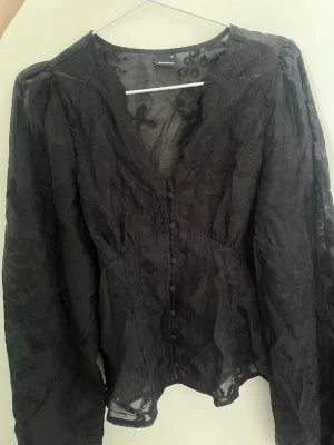 Blus från Gina - Snygg svart blus i transparent spets från Gina med v-ringning och klädda knappar framtill. Blusen har långa puffiga ärmar och ett blommigt mönster i tyget. 