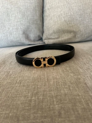 Svart skärp från Salvatore Ferragamo - Snyggt svart skärp från Salvatore Ferragamo med ikoniskt guldigt spänne i metall. Bältet är gjort i slätt skinn och har en stilren, klassisk design som passar till många olika outfits.