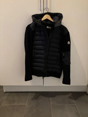 Moncler cardigan - Säljer en svart pufferjacka från Moncler med quiltad front och huva. Jackan har en cool patch på ärmen och dragkedja fram. Materialet är en mix av dun och stickad textil, vilket ger både värme och stil. Perfekt för kalla vinterdagar.