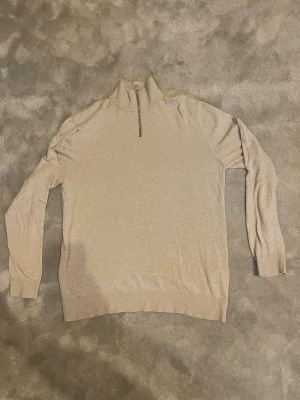 Beige stickad halv zip tröja från Jack & Jones  - Snygg beige stickad tröja med hög krage och halv dragkedja framtill. Tröjan har långa ärmar och ribbade muddar vid ärmslut och nederkant. Perfekt för lager på lager och enkel att matcha med olika outfits.