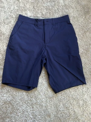Mörkblå golf shorts från Lyle & Scott - Snygga mörkblå golf shorts från Lyle & Scott med klassisk passform och diskret logga i gult bak. Shortsen har sidofickor, bälteshällor och är tillverkade i ett lätt och bekvämt material, perfekt för varma dagar.
