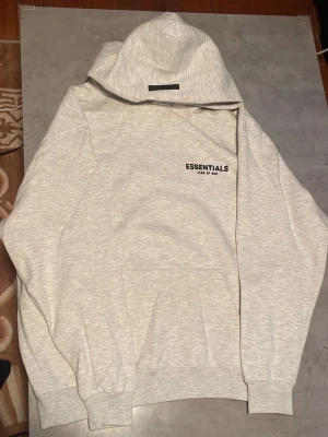 Essentials Fear of God grå hoodie - Ljusgrå hoodie från Essentials Fear of God med svart logga tryckt på bröstet och ryggen. Klassisk passform med huva och ribbade muddar vid ärmar och midja. Tillverkad i mjukt bomullsmaterial som känns skönt mot huden. Perfekt för en clean och avslappnad stil.