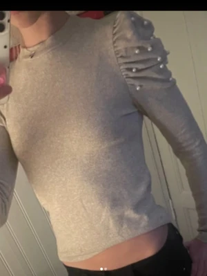 Beige topp med puffärm och pärlor - Trendig beige topp med långa puffärmar dekorerade med pärlor. Toppen har en slim passform och rund halsringning. Perfekt för dig som vill ha något extra till din outfit.
