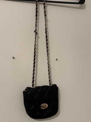 Svart quiltad axelväska med kedja - Snygg svart axelväska i quiltat material med guldfärgad vridknäppe framtill. Väskan har en lång kedjerem med insyddt svart band, perfekt för att bära crossbody. Liten och smidig modell som rymmer det viktigaste. Passar till många olika outfits.