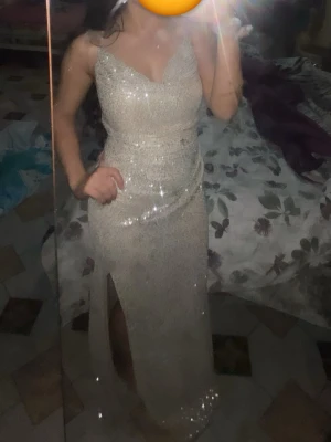 Glittrig långklänning med slits - Supersnygg glittrig långklänning i ljus champagnefärg med smala axelband och v-ringning. Klänningen har en hög slits framtill och sitter figurnära för en elegant look. Perfekt för dig som vill sticka ut på bal eller fest.